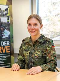 Eine Soldatin sitzt lächelnd an einem Tisch, dahinter ein Roll-up „Dienen in der Reserve“