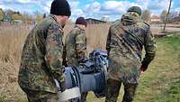 Drei Soldaten in Flecktarnuniform tragen einen Motor über eine Wiese.