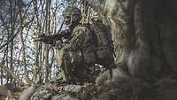 Ein Soldat, der komplett vermummt ist, hockt mit seinem Gewehr vor einem Baum