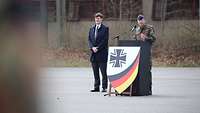 Ein Soldat steht hinter einem Rednerpult. Neben ihm ein Mann in einem Mantel