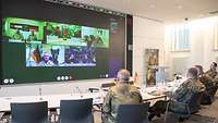 Mehrere Soldaten sitzen an einen Tisch vor einer Videoleindwand auf der Soldaten zu sehen sind.