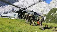 Several soldiers move in single file towards a helicopter on a meadow in front of a steep rock.