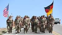 Eine Marschformation mit der deutschen und amerikanischen Flagge geht eine Landstraße entlang.