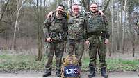 Drei Soldaten Arm in Arm auf einem Waldweg. Vor ihnen steht ein Militärrucksack, auf dem „13K3“ steht. 