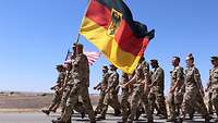 Eine Marschformation mit der deutschen und amerikanischen Flagge geht eine Landstraße entlang