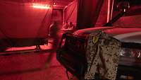 Drei Soldaten sitzen unter einer roten Lampe, im Vordergrund hängt eine Jacke mit einem MAD-Aufnäher auf einem Geländewagen.