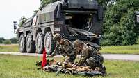 Zwei Soldaten versorgen eine verletzte Person, im Hintergrund steht ein Radpanzer mit rotem Kreuz.