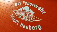 Das Wappen der Bundeswehr-Feuerwehr Heuberg weiß auf roter Ziegelwand gemalt