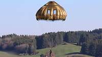 Ein Soldat mit goldglänzendem Schirm schwebt über einer grünen Wiese Richtung Boden.