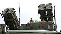 Ein Soldat steht auf dem Führerhaus eines Lkw, auf dem sich Flugabwehrraketen befinden, die in Richtung Himmel zeigen. 