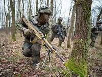 Ein Soldat mit Maschinengewehr rennt durch den Wald. Weitere Soldaten im Hintergrund.