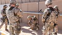 Ein Soldat mit Helm, Weste und Waffe erklärt Soldatinnen und Soldaten das weitere Vorgehen, in dem er am Boden Zeichen aufmalt.