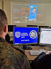 Zwei Männer sitzen vor zwei Monitoren mit der Inschrift Locked Shields 2023 Blue Team 03.