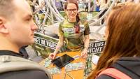 Frau an Tisch mit Infomaterial. Davor ein Mann und eine Frau. Der Mann blickt zur Frau.