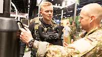 Ein Soldat spricht mit jungem Mann, der ein Tauchgerät trägt. Der Soldat fasst an ein anderes Gerät.