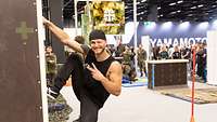 Kräftiger junger Mann im Sportanzug hängt einhändig an Kasten, im Hintergrund der Bundeswehr-Stand.