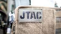 Auf einer beigen Klettfläche stehen auf einem beigen Patch in Schwarz die Buchstaben JTAC.