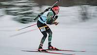Eine Biathletin skatet mit dem Gewehr auf dem Rücken von links kommend. 