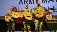 Drei Soldaten der U.S. Army National Guard stehen nebeneinander. Sie halten jeweils zwei Trophäen.