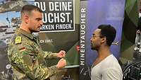 Ein Mann in Uniform spricht mit einem Besucher ihm gegenüber. Dahinter Gerät und Poster.