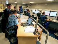 Header Leitstand F222 Mehrere Personen und blauer Arbeitsuniform sitzen verteilt an zwei langen Computerarbeitsplatzreihen
