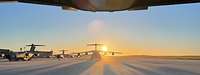Zu sehen sind mehrere Transportflugzeuge auf einem Rollfeld, mit untergehende Sonne im Hintergrund.