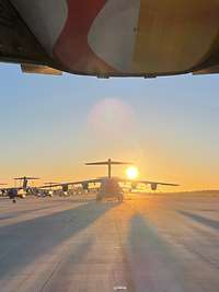 Zu sehen sind mehrere Transportflugzeuge auf einem Rollfeld, mit untergehende Sonne im Hintergrund.