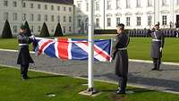 Zwei Soldaten falten eine Flagge wärend die anderen Soldaten salutieren mit Blick zur Flagge. 
