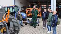 Zwei Jungen und drei Mädchen stehen auf einem Technikhof. Ein Geländebetreuer zeigt verschiedene Kfz