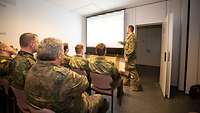 Ein Soldat steht an einer Beamerleinwand vor sitzenden Soldaten und hält einen Vortrag.