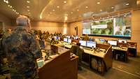 Mehrere Soldaten stehen und sitzen an Computern in einem Raum vor einem großen Wanddisplay.