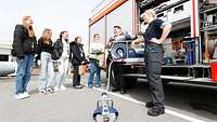 Eine Feuerwehrfrau und ein Feuerwehrmann erklären einer Gruppe Mädchen die Ausstattung des Fahrzeugs