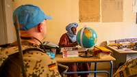 Begrüßung durch den Schuldirektor Ein Mann mit Turban sitzt an einem Tisch, auf dem ein Globus steht. Im Vordergrund sind zwei Soldaten von hinten zu sehen.