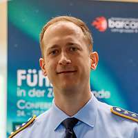 Hauptmann Michael Z. blickt in die Kamera, hinter ihm ist ein Plakat des „BarCamps“ zu sehen