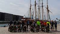 Fahrradfahrer stehen für ein Gruppenbild in einem Hafen zusammen. Im Hintergrund steht ein Segelschiff.