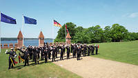 Musiker in Uniform stehen auf einer Wiese vor mehreren Flaggen