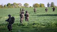 Bewaffnete Soldaten laufen in Abständen nebeneinander über ein grünes Feld in der Sonne.