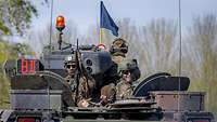 Drei Soldaten schauen aus den Luken eines fahrenden Panzers mit blauem Wimpel und Rundumleuchte.