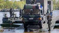 Zwei Lastwagen verlassen eine Fähre aus Amphibienfahrzeugen. Daneben stehen Soldaten.