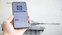 Auf einem Smartphone ist eine Anmeldemaske abgebildet, im Hintergrund ein Panzer auf einem Display