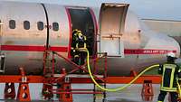 Feuerwehrleute klettern in ein Flugzeug, ein Feuerwehrmann hält einen Löschschlauch. 