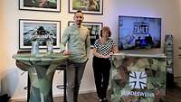 Ein Mann und eine Frau an Stehtischen mit Bundeswehr-Logo. Dahinter Bilder und ein Fernseher.
