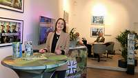 Eine Frau steht an einem Tisch, dahinter eine Lounge mit Sitzecke und Bundeswehrpos-tern.