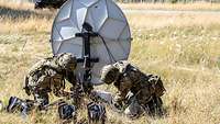Zwei Soldaten arbeiten an einer Satellitenanlage auf einer trockenen Wiese.