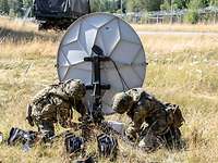 Zwei Soldaten arbeiten an einer Satellitenanlage auf einer trockenen Wiese.