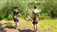 Bundeswehr Olympix 2023 Ein Mann schwingt an einer Seilbahn über einen Graben. Zwei weitere erwarten ihn am anderen Ufer.