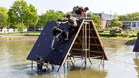Bundeswehr Olympix 2023 Vier Männer klettern über ein Hindernis. Einer steht in hüfthohem Wasser. Die anderen sind auf dem Hindernis.