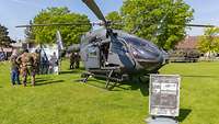Ein Helikopter auf einer Wiese, davor ein Infoschild, daneben Betrachtende.