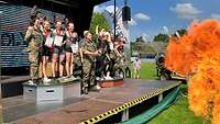 Bundeswehr Olympix 2023 Siegerehrung, Gruppen junger Frauen auf einem Podest, im Vordergrund eine Flamme.