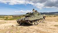 Ein Kampfpanzer fährt durch kahles Gelände, ein Soldat schaut mit einem Fernglas aus der Luke.